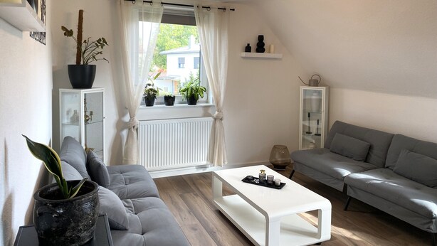 A modern living room with a grey sofa, a white coffee table, and a window with curtains. There are potted plants on the windowsill and a white cabinet with glass doors. - Modern feel-good apartment in the Illertal (Dettingen an der Iller)