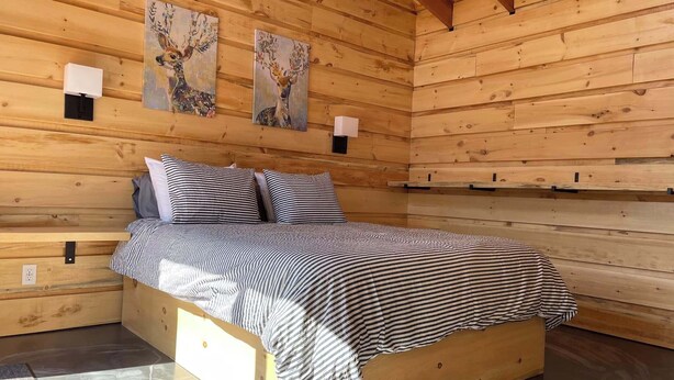 A wooden cabin bedroom with a bed, two wall-mounted lamps, and two paintings of deer. - Chalet L'Orée Étoilée (La Bostonnais)