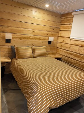 Een kamer met een wand bekleed met houtpaneel, een bed met gestreept beddengoed, twee nachtlampjes en een raam met een zonnescherm.