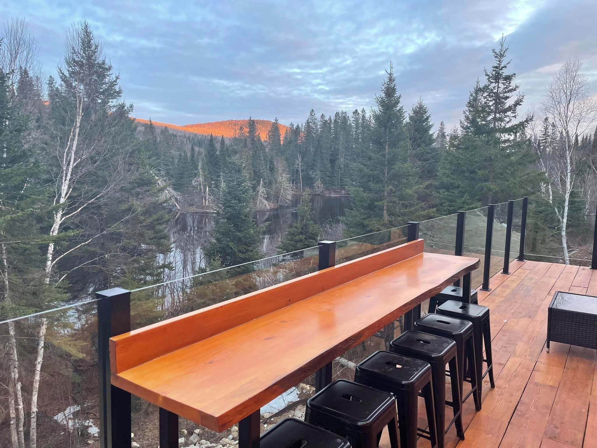 A wooden deck with a bench and black stools, overlooking a forested area with a lake and distant hills.