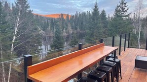 A wooden deck with a bench and black stools, overlooking a forested area with a lake and distant hills. - Chalet L'Orée Étoilée (La Bostonnais)