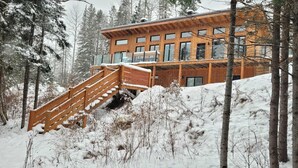 A wooden cabin with a staircase leading to a deck, surrounded by snow-covered trees. - Chalet L'Orée Étoilée (La Bostonnais)