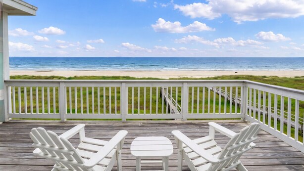 A wooden deck with white chairs and a table, overlooking a beach with a clear sky. - Beachfront | 4BR | Sleeps 14 | Pet-Friendly | NEW Hot Tub (Galveston)
