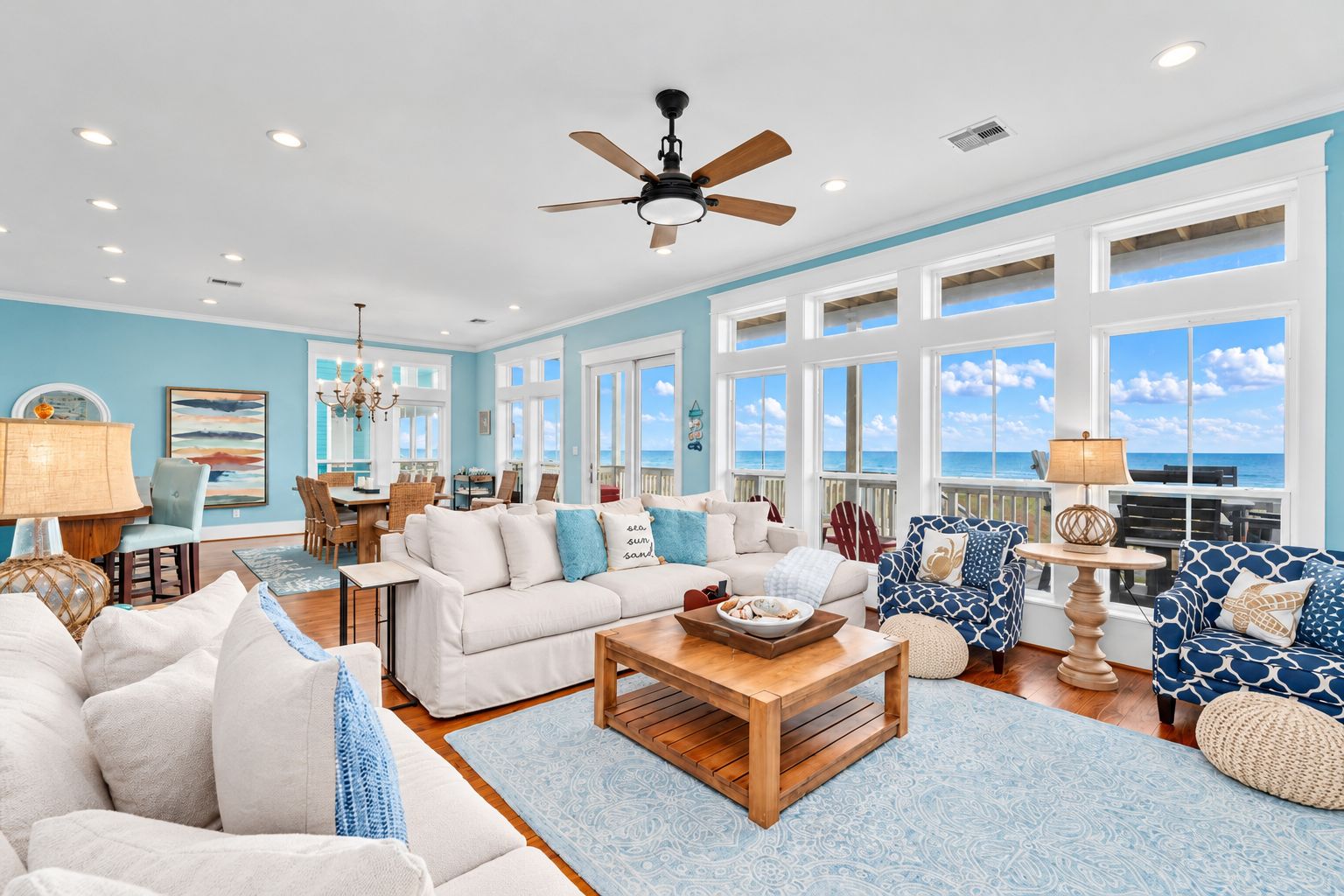 A spacious living room with a large window overlooking the ocean, a ceiling fan, and a dining area with a view.