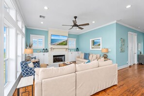 A living room with a beige sectional sofa, a fireplace, a ceiling fan, and a large TV mounted above it. - Beachfront | 4BR | Sleeps 14 | Pet-Friendly | NEW Hot Tub (Galveston)