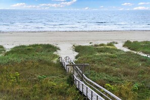 Une promenade en bois mène à une plage bordée de dunes herbeuses, avec l’océan en toile de fond.