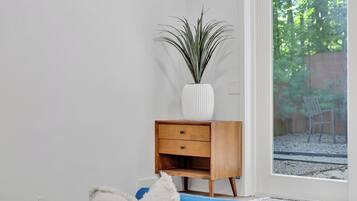 Ein Zimmer mit Nachttisch aus Holz, einer weißen Vase mit Pflanze und Blick auf einen Garten durch eine Glastür.