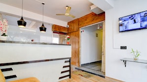 A modern reception area with a wooden reception desk, a sofa, pendant lights, a ceiling fan, and a mounted television. - Hotel Casa Inn (Baruipur)