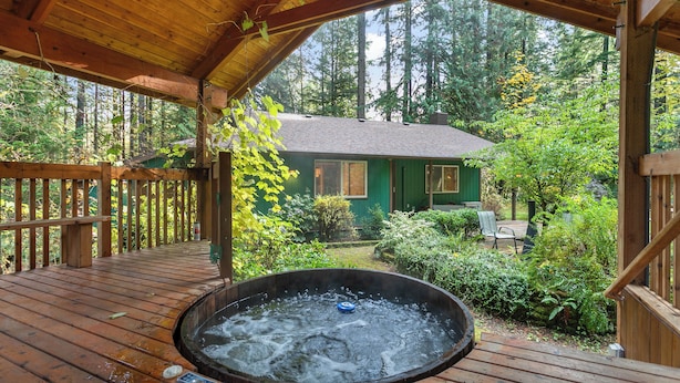 Una terraza de madera con un jacuzzi, rodeada de exuberante vegetación y una cabaña al fondo.