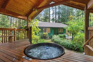 Una terraza de madera con un jacuzzi, rodeada de exuberante vegetación y una cabaña al fondo.