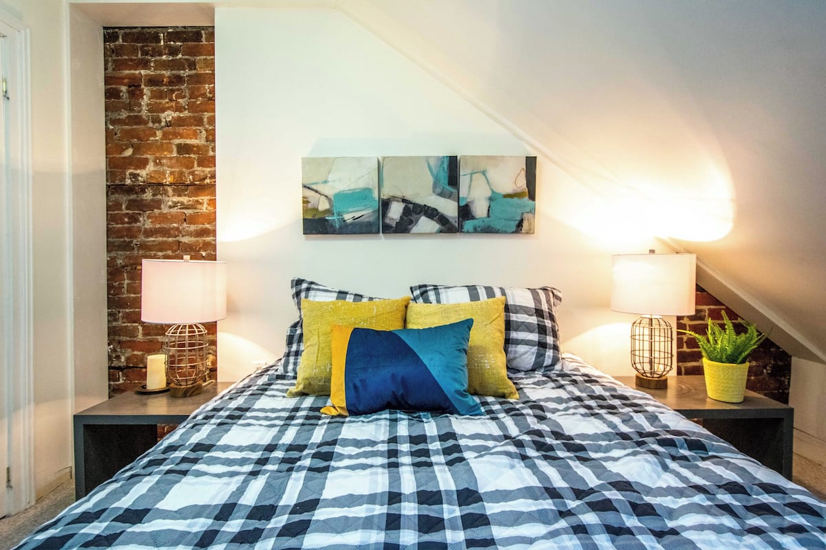 A bedroom with a checkered bedspread, two bedside lamps, a potted plant, and a brick wall.