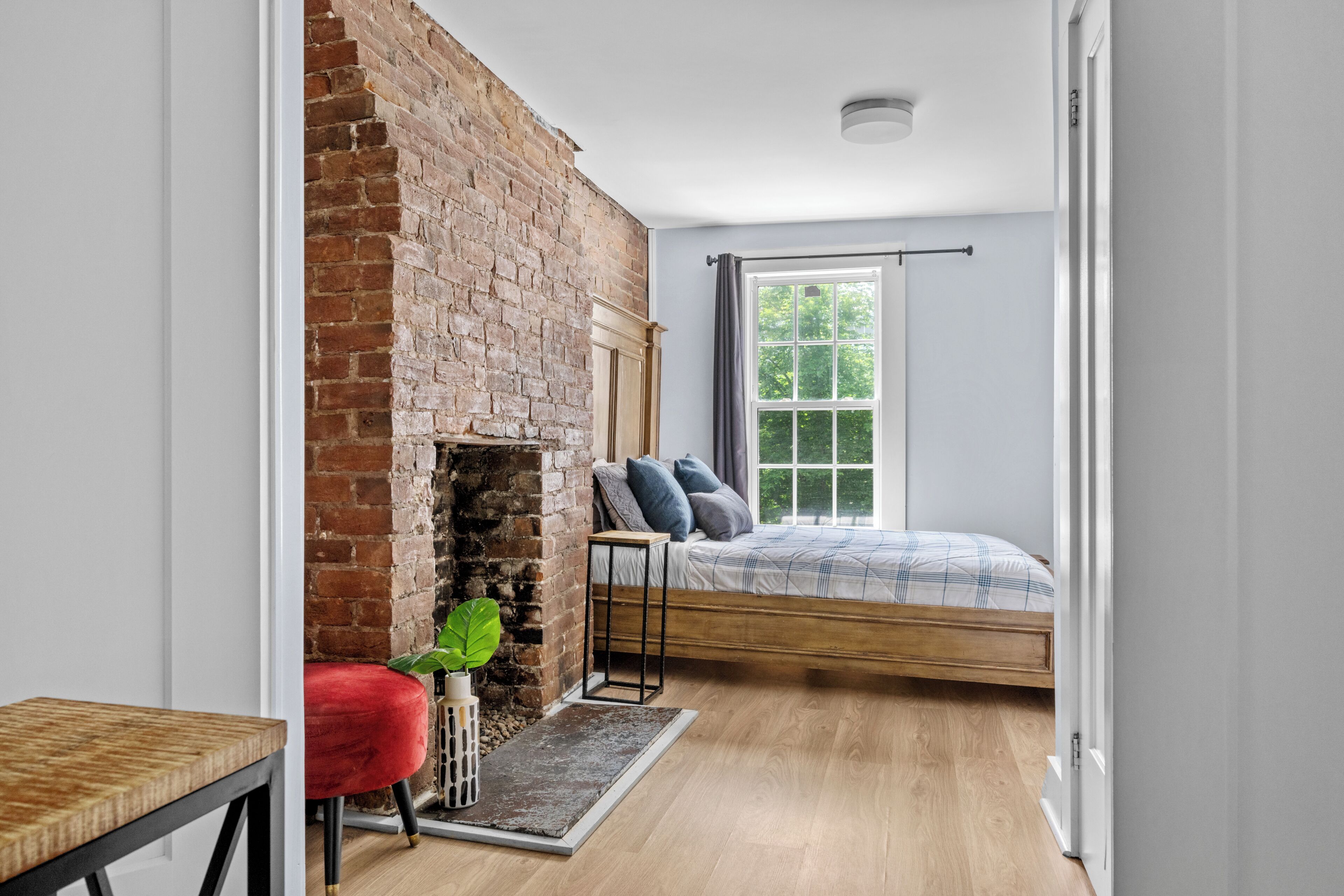 Un dormitorio con chimenea de ladrillo, cama con cubrecama escocés, una silla roja y una mesita.