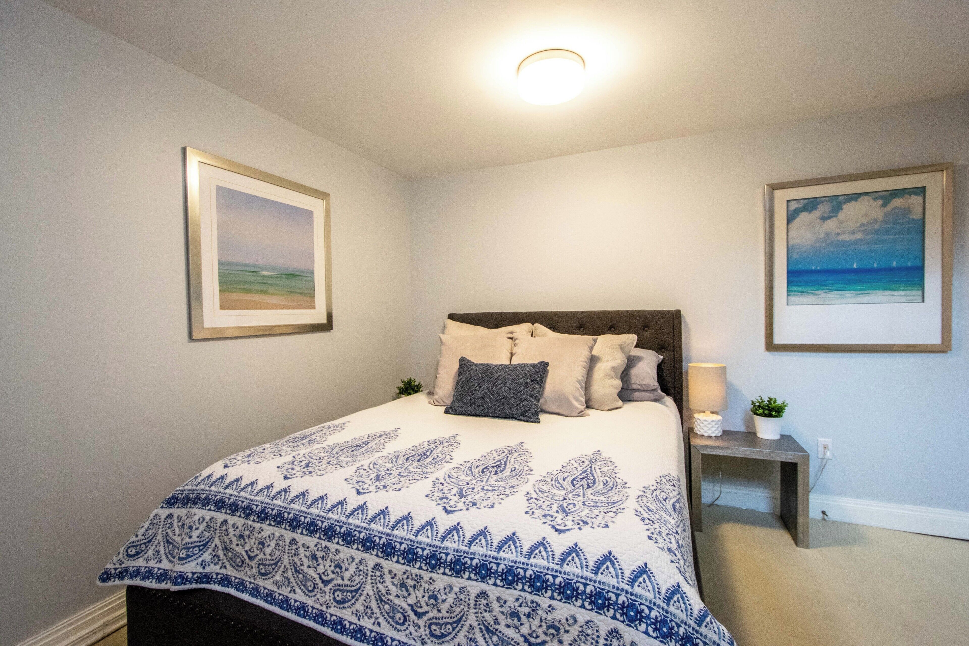 A bedroom with a bed, pillows, a bedside table with a lamp, and two framed pictures on the wall.