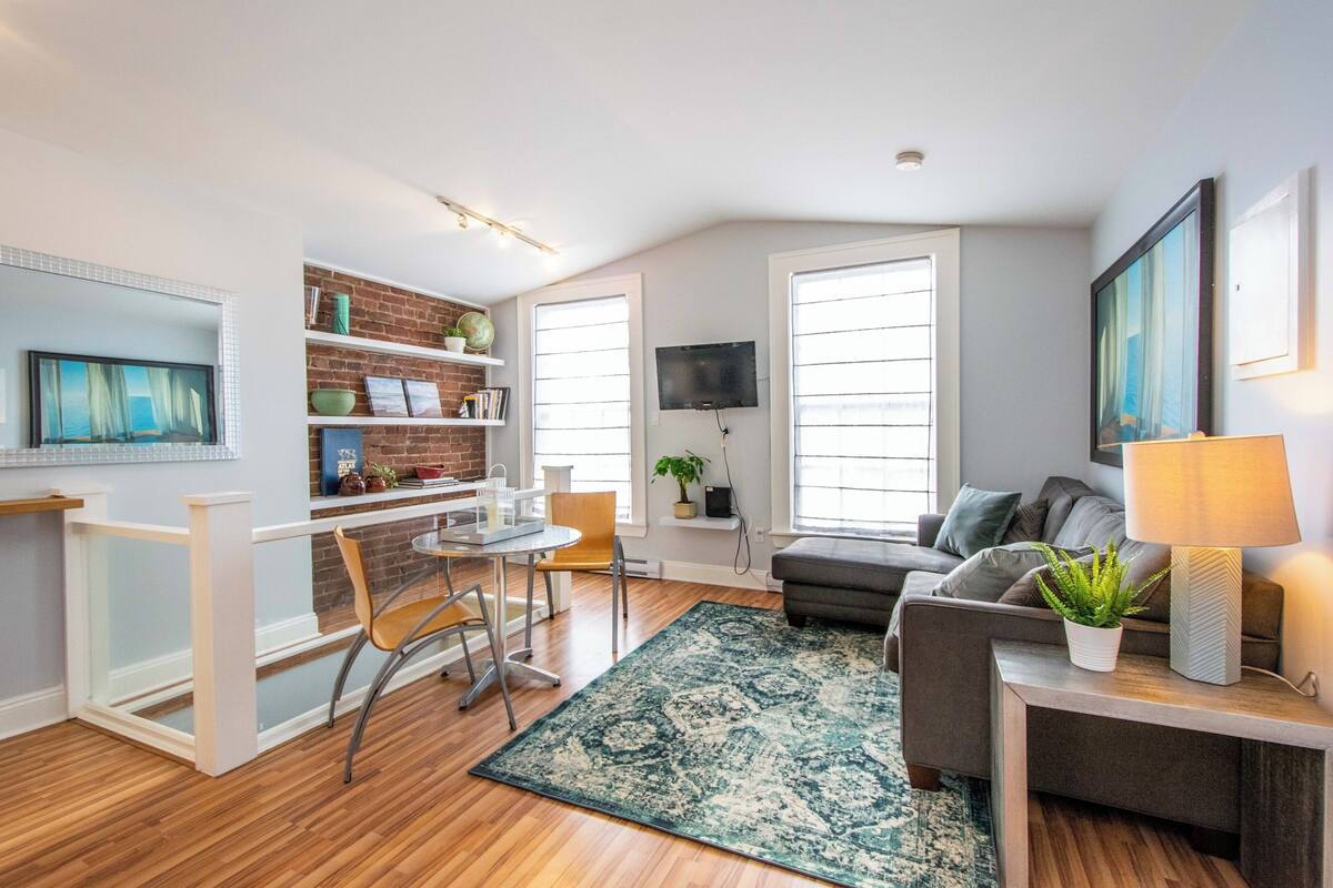 A modern living room with a dining area, a grey sofa, a TV mounted on the wall, and a bookshelf with decorative items.