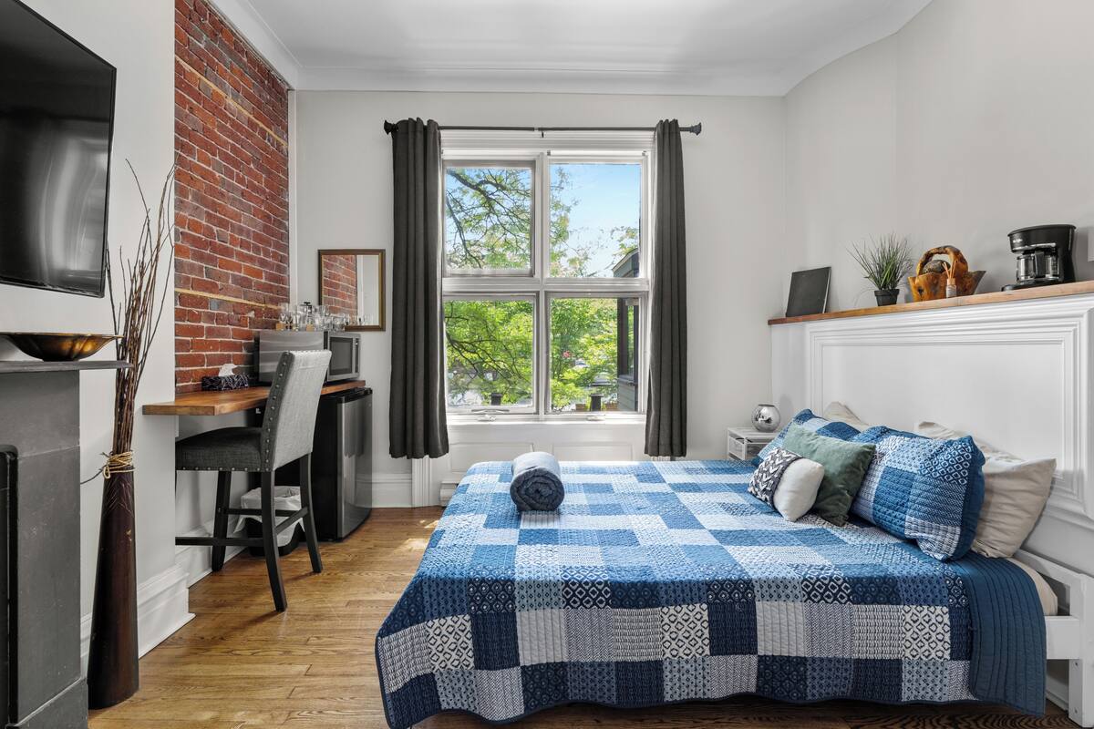 A compact bedroom with a checkered bedspread, a desk with a chair, a television, and a brick accent wall.