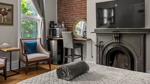A bedroom with a brick wall, a fireplace, a TV, a desk with a chair, a bed with a blanket, and a window with curtains. - Red George Inn and Suites (Halifax)