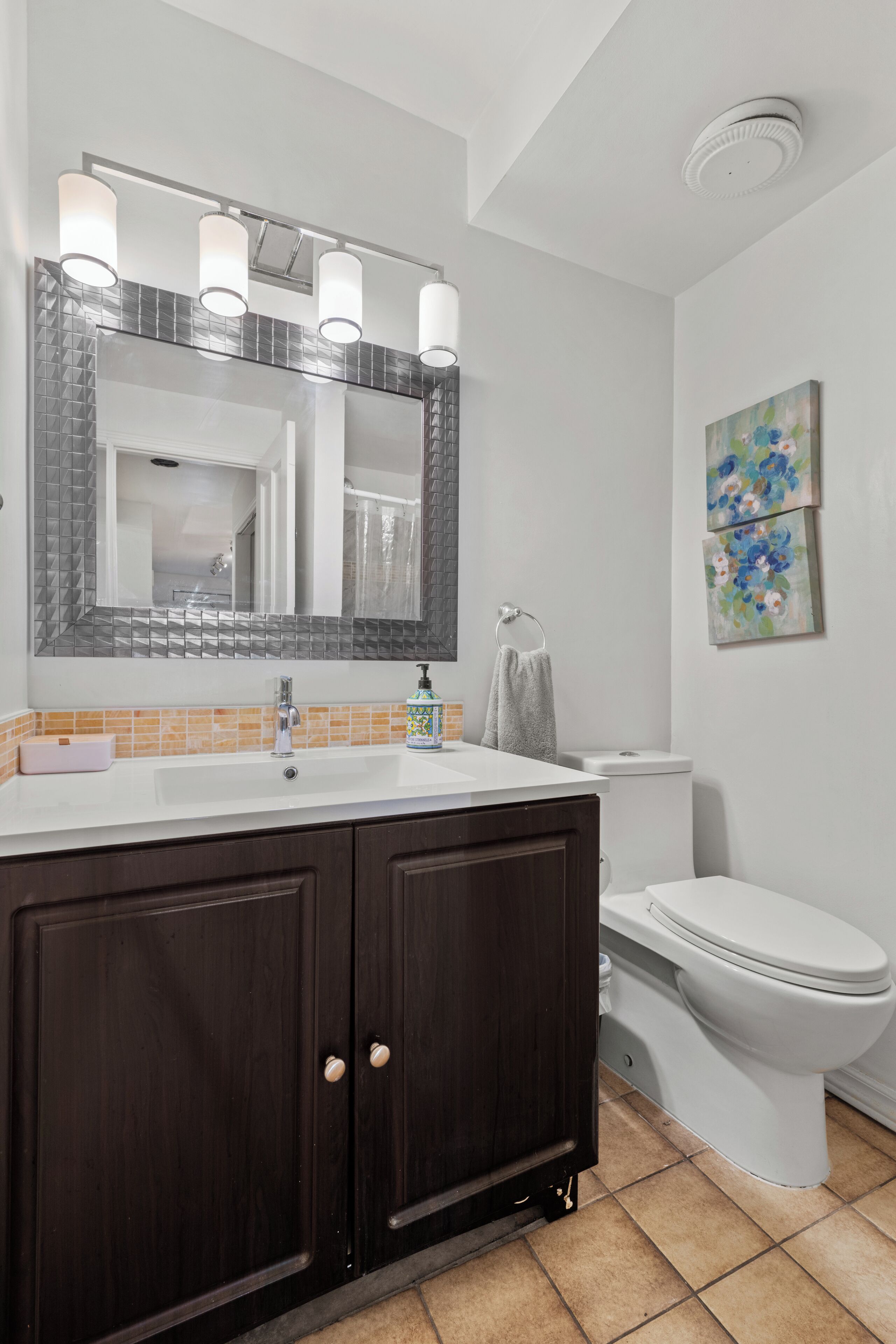 A bathroom with a white sink, dark wooden cabinets, a large mirror with lights, a toilet, and a small painting on the wall.