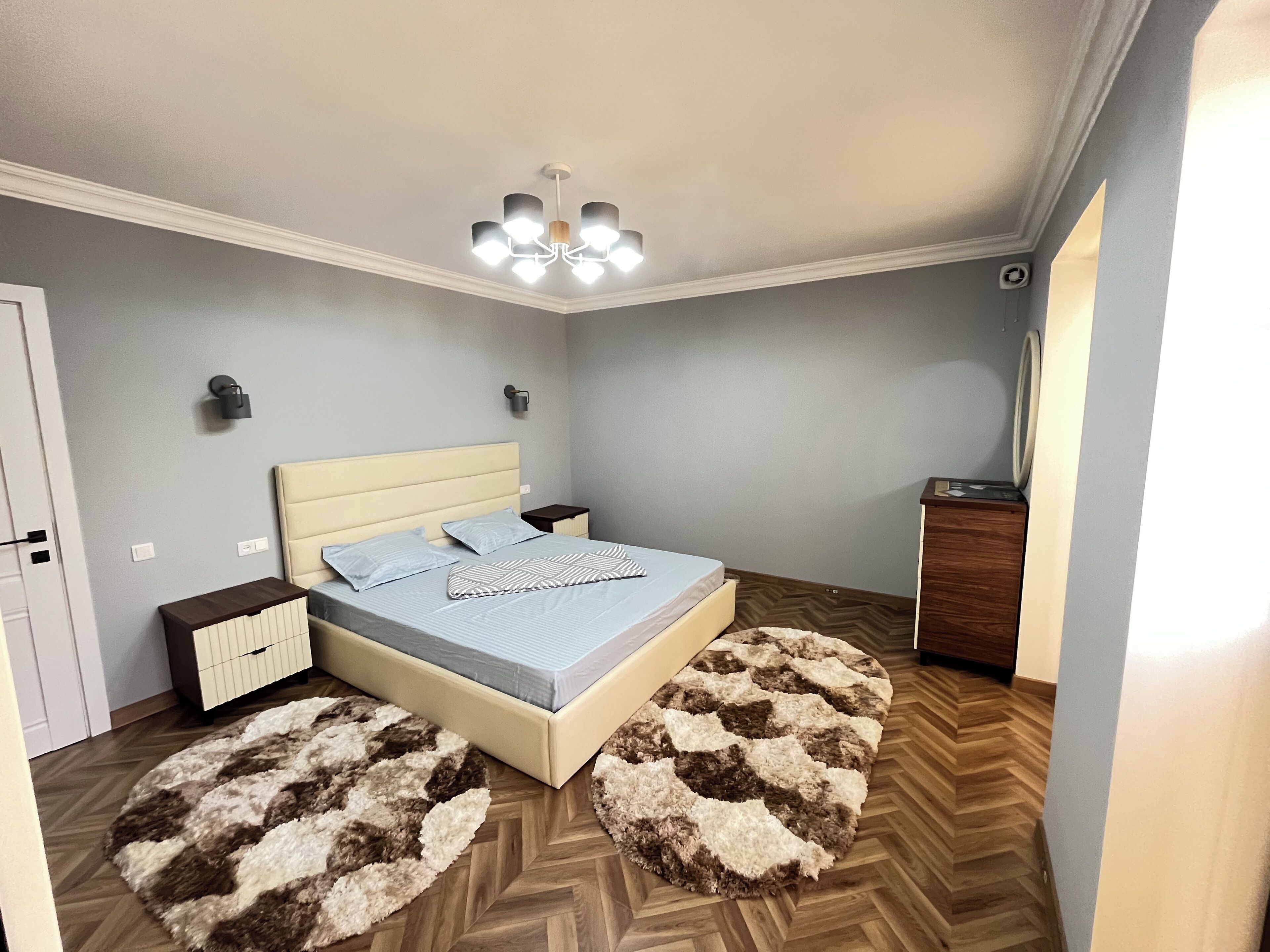 A bedroom with a bed, bedside tables, a dresser, a ceiling light fixture, and a herringbone-patterned wooden floor.