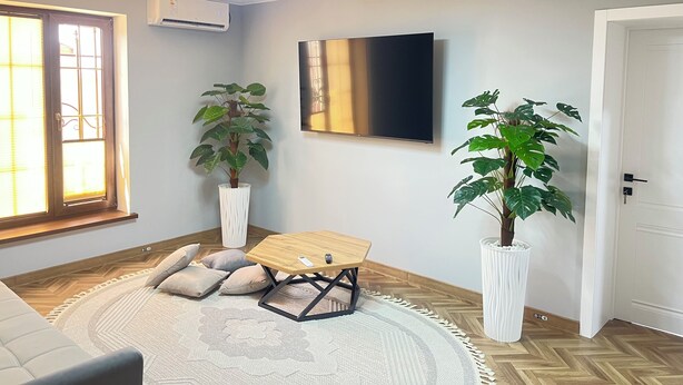 Een moderne woonkamer met een houten salontafel, twee potplanten, een flatscreen televisie en een groot raam met zonwering.