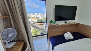 A hotel room with a bed, a TV mounted on the wall, a fan, and a view of the cityscape. - COSTE0109 - Concept Life Stella (Salvador)