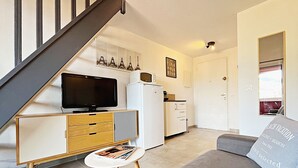 A modern living room with a sofa, a television, a small table with magazines, a white refrigerator, and a staircase. - Apartment 6 people sea view - Théoule sur mer (Côte d'Azur) - SAVANNAH (Théoule-sur-Mer)