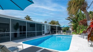 En swimmingpool med liggestole og parasoller, omgivet af en bygning med store vinduer og en overdækket terrasse.