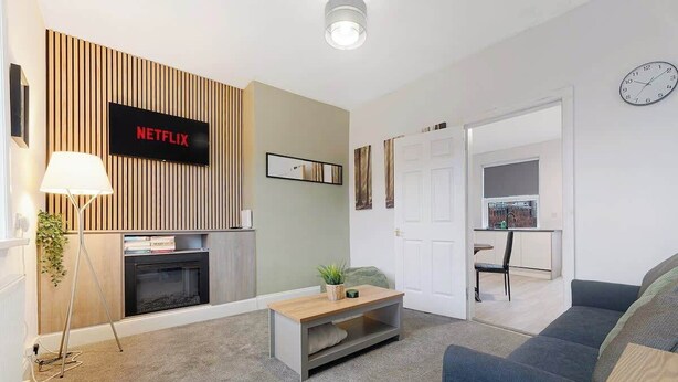 Ein modernes Wohnzimmer mit Kamin, Flachbildfernseher, einer Couch und einem Essbereich, der durch eine offene Tür sichtbar ist.