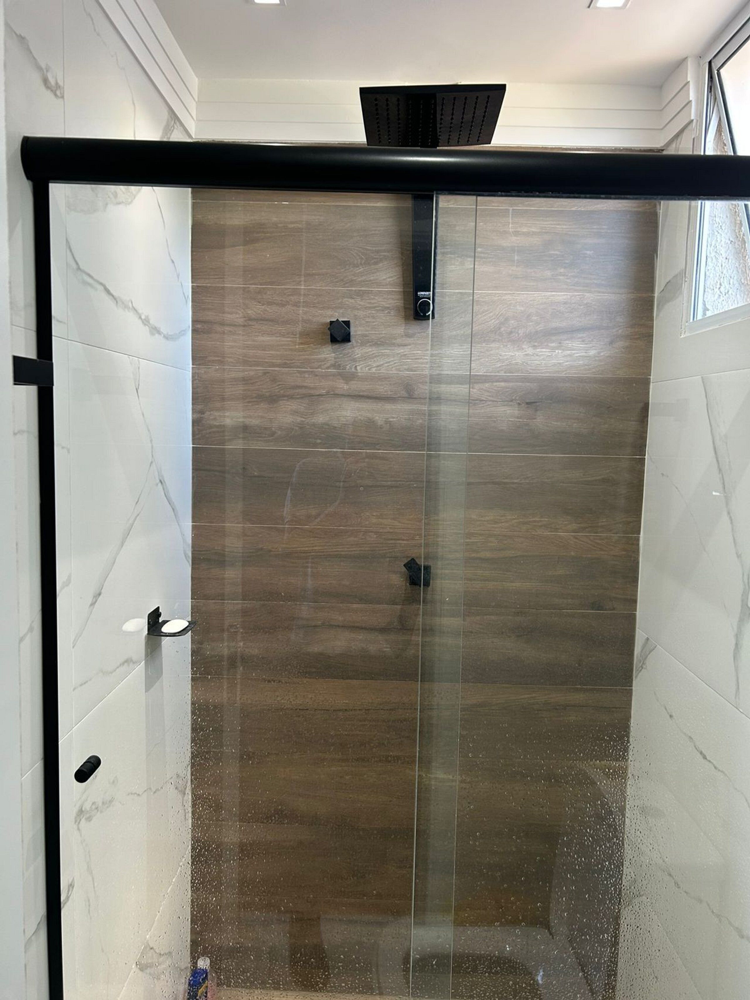 Cabina doccia in vetro con parete in pannelli di legno, un pannello comandi nero e una finestra sulla destra.