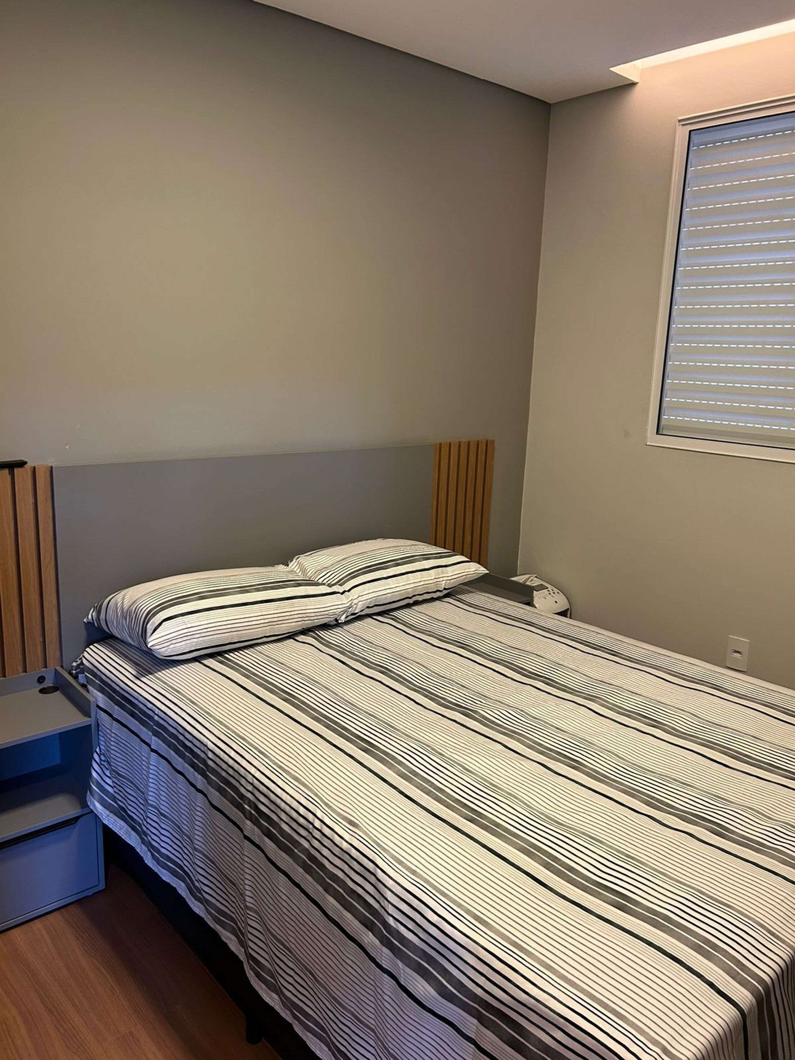 A bedroom with a bed, a nightstand, a wooden headboard, and a window with blinds.