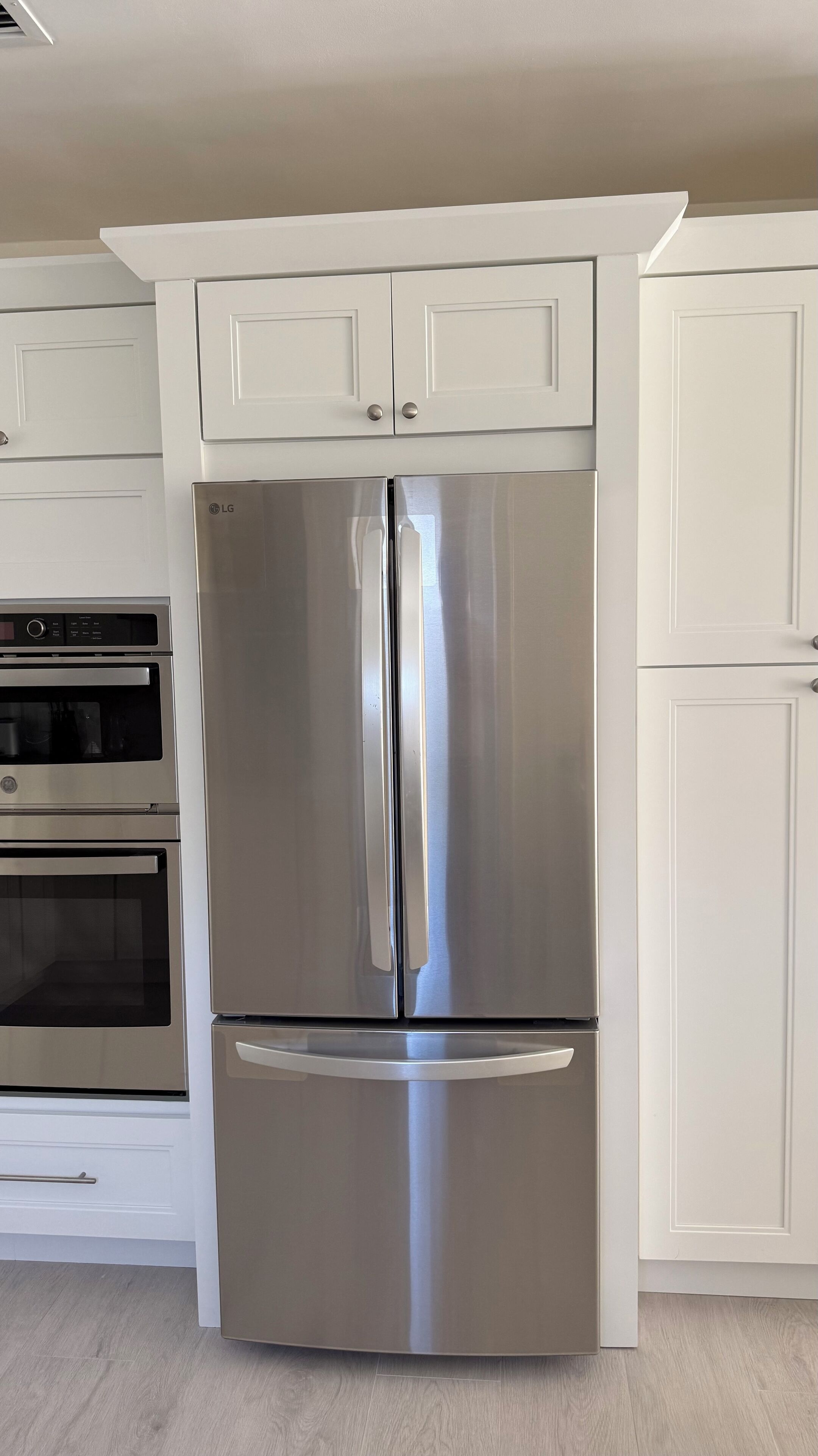 A stainless steel refrigerator with a built-in oven, white cabinetry, and a light-colored wooden floor.