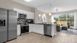 A modern kitchen with stainless steel appliances, white cabinets, and a large island. There is a dining area with a gray sofa and a view of the outdoors. - 3 Bedrooms / 3 Bathrooms / Windsor Hills (7662 SR) (Kissimmee)
