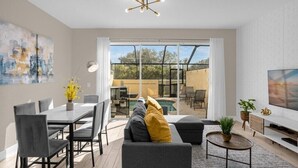 A modern living room with a dining area, a sofa with cushions, a TV, and a large window with a view of the outdoors. - 3 Bedrooms / 3 Bathrooms / Windsor Hills (7662 SR) (Kissimmee)
