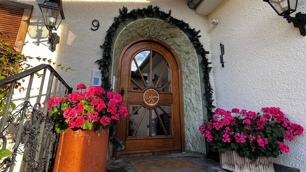 A wooden door with a circular emblem, flanked by two large flower pots with pink flowers, and a stone archway above the door. - Fackler Apartments 1 Riederstein - Relaxed Bavarian Living Near Lake Tegernsee (Tegernsee)