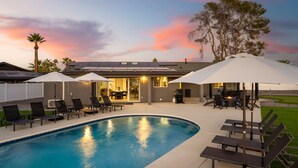Una piscina con sillas de descanso y sombrillas, un porche cubierto y una casa con puerta de vidrio.