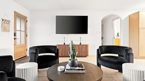 Una sala moderna con una mesa redonda de madera, dos sillones negros y un reposapiés blanco. Un televisor de pantalla plana está montado en la pared sobre la mesa.