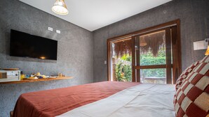 Un dormitorio con una cama grande, un televisor de pantalla plana montado en la pared, una repisa de madera con artículos de desayuno y un ventanal con vistas a la vegetación.