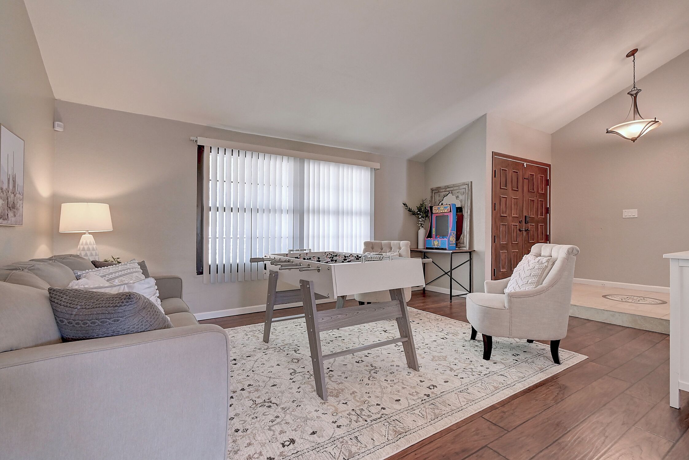 A living room with a sofa, a coffee table, a foosball table, a chair, and a window with blinds.