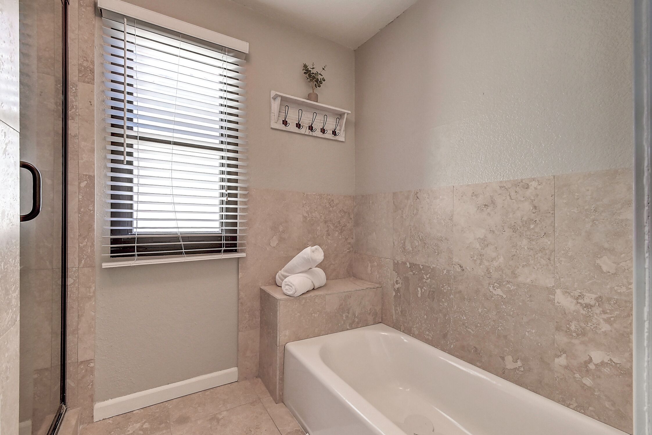 A bathroom with a bathtub, a window with blinds, a towel rack, and a shelf with towels.