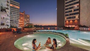 Area piscina sul tetto con tre persone che si rilassano in acqua, circondata da edifici e un cielo notturno.