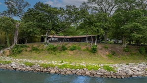 A lakeside cabin with a covered patio, surrounded by trees and a rocky shoreline.