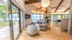 A spacious living room with a large window, a round ottoman, and a dining area with a view of the outdoors.