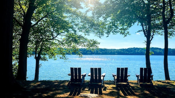 A lakeside view with four Adirondack chairs facing the water, surrounded by trees.