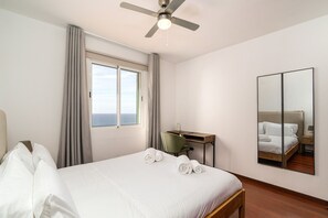 A hotel room with a bed, a desk, a chair, a ceiling fan, a window with curtains, and a mirror on the wall. - Mediterranean Style • Sound of Waves • Sunsets (Arco da Calheta)