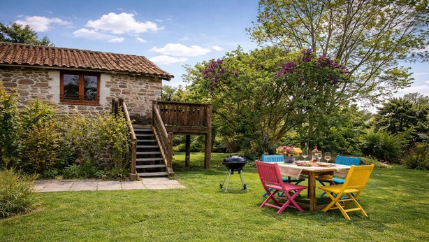 A stone house with a wooden staircase leading to a deck, surrounded by lush greenery and a dining area with colorful chairs.