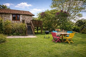 Une maison en pierre avec un escalier en bois menant à une terrasse, entourée d’une végétation luxuriante et dotée d’un coin repas avec des chaises colorées.