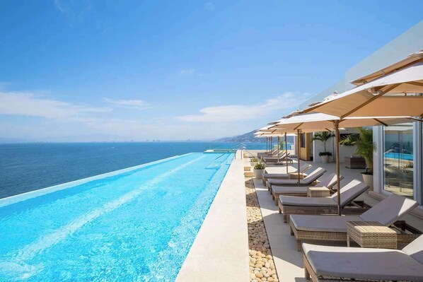 Un espace aménagé au bord de la piscine, avec des chaises longues et des parasols, offrant une vue sur un océan d’un bleu limpide.