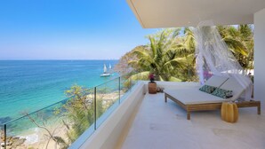 Un balcon avec un fauteuil de relaxation blanc doté de coussins, un pot de fleurs et une vue sur la mer, où l’on aperçoit des voiliers.