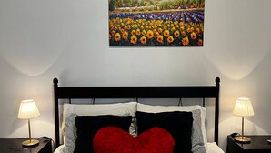 A hotel room with a bed, a red heart-shaped pillow, two bedside tables with lamps, an air conditioner, and a painting of a sunflower field on the wall. - Charming apartment 5' walk to the beach (Carolina)