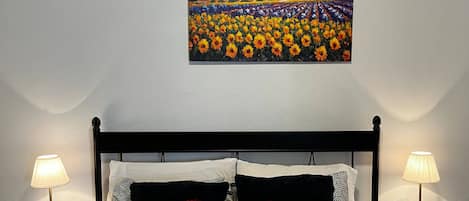 Hotelkamer met een bed, een rood hartvormig kussen, twee nachtkastjes met lampen, een airconditioner en een schilderij van een zonnebloemenveld aan de muur.