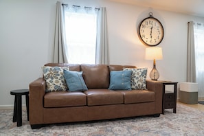 A brown leather sofa with patterned cushions, a small side table with a lamp, and a wall clock.